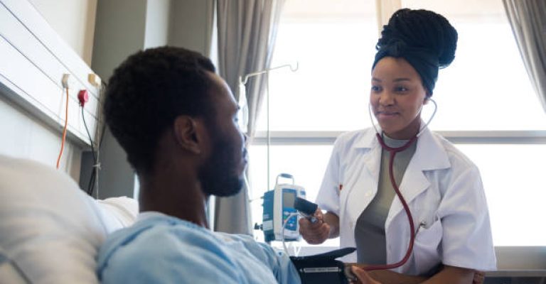 Smiling female doctor looking at male patient while checking blood pressure. Confident healthcare worker is standing with man in hospital ward. They are in medical clinic.